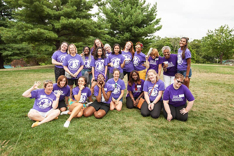 Group of Big Sister Boston staff members all wearing purple shirts and posing together for a group photo on a grassy field.